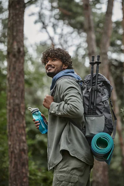 Позитивный молодой индийский путешественник держит рюкзак и спортивную бутылку во время прогулки в лесу — стоковое фото