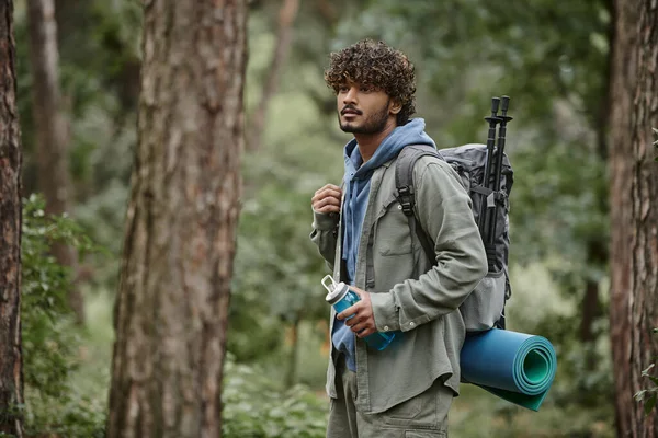 Encaracolado jovem turista indiano em camisa segurando mochila e garrafa de esportes na floresta — Fotografia de Stock
