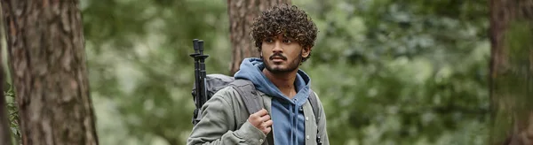 Молодий кучерявий індійський турист з рюкзаком і пішохідними полюсами, дивлячись в ліс, банер — стокове фото