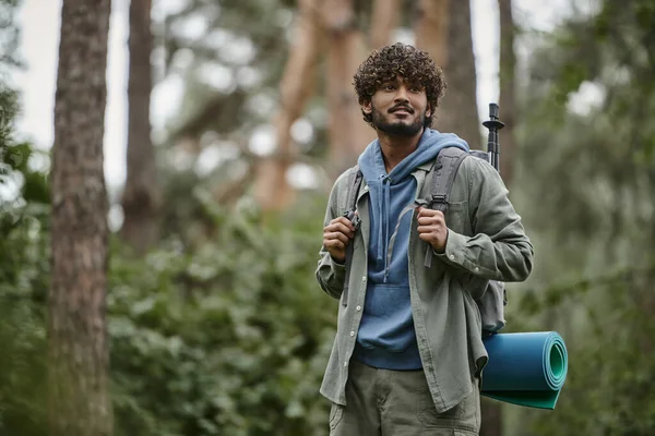 Joven viajero indio contento sosteniendo la mochila y mirando hacia otro lado en el bosque - foto de stock