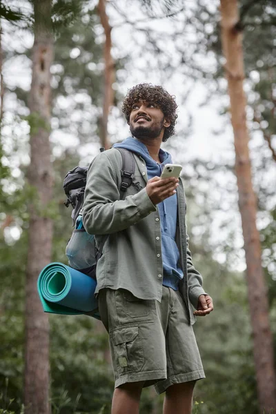Low angle view of cheerful young indian tourist with backpack using smartphone in forest — Stock Photo