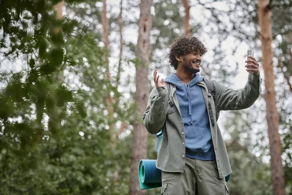 Позитивный молодой индийский турист с рюкзаком с видеозвонком на мобильный телефон в лесу — стоковое фото