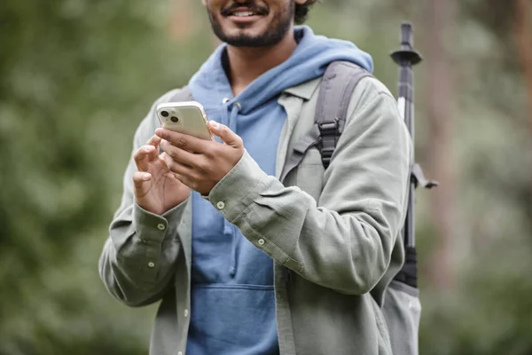 Cropped view of smiling indian hiker with backpack using smartphone in blurred forest — Stock Photo