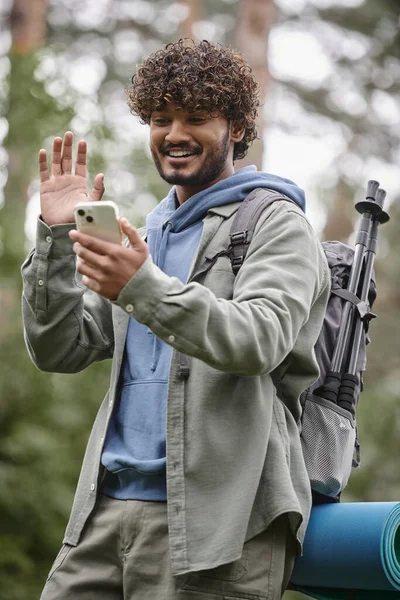 Overjoyed indian hiker with backpack waving hand while having video call on smartphone in forest — Stock Photo