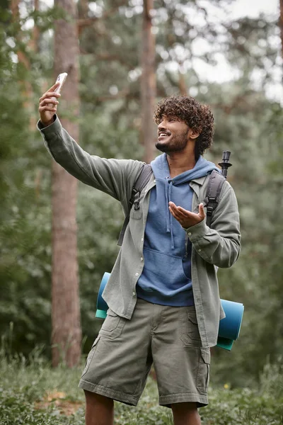 Positiver junger indischer Reisender mit Rucksack mit Videotelefon im verschwommenen Wald — Stockfoto