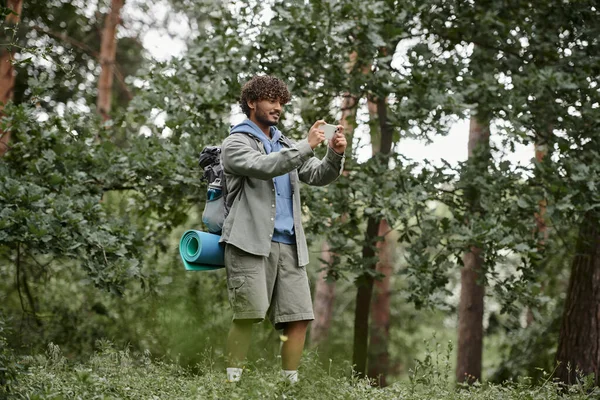 Улыбающийся молодой индийский турист с рюкзаком делает фото на смартфоне в лесу — стоковое фото