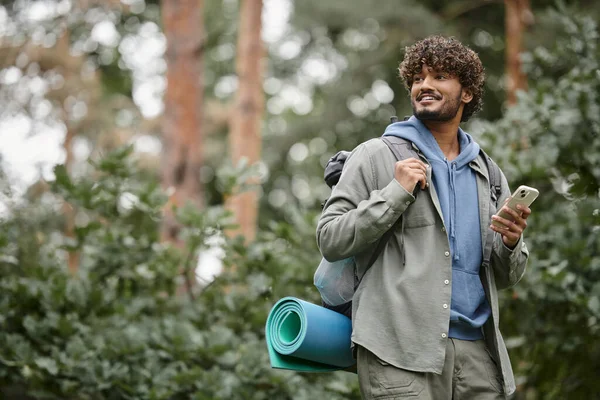 Carefree young indian hiker with backpack holding smartphone and looking away in forest — Stock Photo