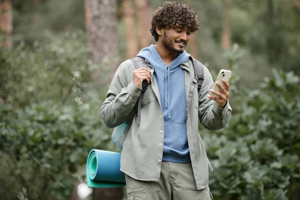 Молодой улыбающийся индийский турист с рюкзаком, используя смартфон, стоя в размытом лесу — стоковое фото