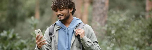 Позитивный молодой индийский турист с рюкзаком, используя смартфон в лесу, баннер — стоковое фото