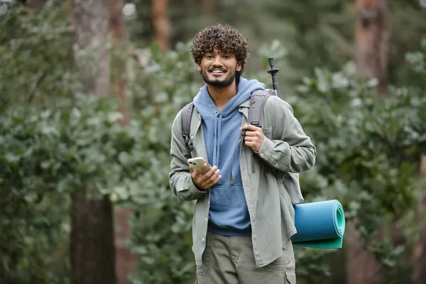 Sonriente joven senderista indio con mochila sosteniendo teléfono inteligente y mirando a la cámara en el bosque - foto de stock