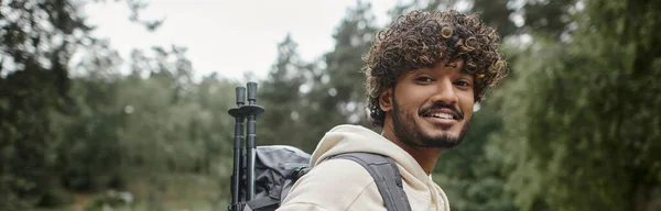 Positive young indian traveler with trekking poles and backpack standing in forest, banner — Stock Photo