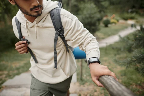 Vista recortada de un joven turista indio con mochila y reloj inteligente caminando cerca de la cerca en el bosque - foto de stock
