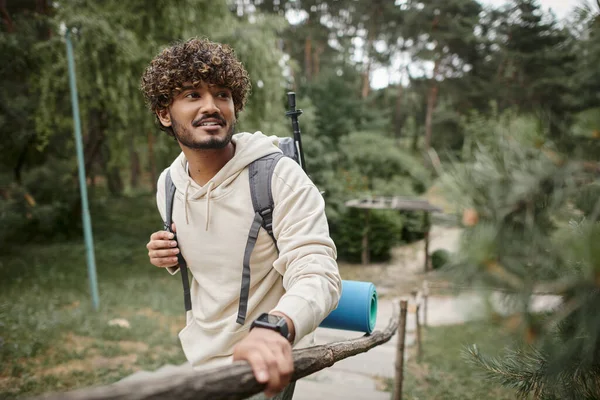 Turista indio sonriente con mochila y reloj inteligente de pie cerca de valla en el bosque - foto de stock