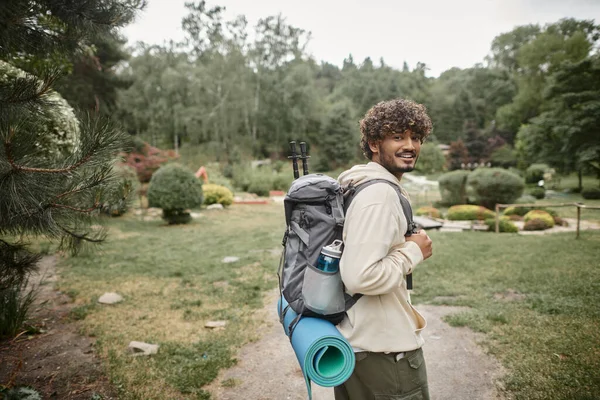 Улыбающийся молодой индийский турист с рюкзаком и пешеходными палками, смотрящий на камеру в лесу — стоковое фото