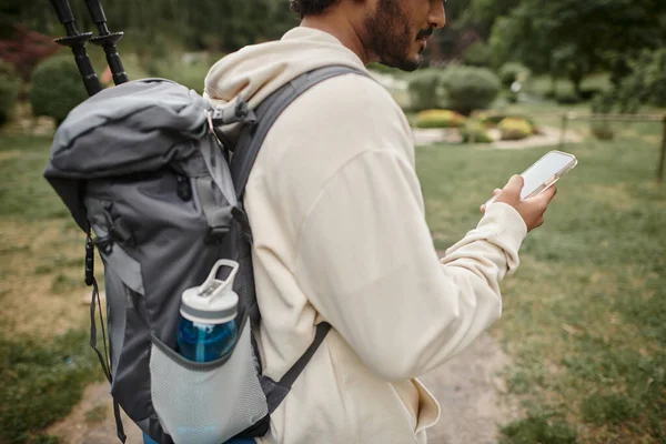 Cropped view of indian hiker with backpack using smartphone on path in forest — Stock Photo