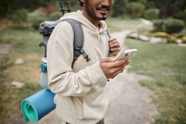 Cropped view of smiling indian traveler with backpack using smartphone in forest — Stock Photo