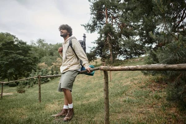 Беззаботный индийский турист с рюкзаком, стоящим возле деревянного забора в лесу — стоковое фото