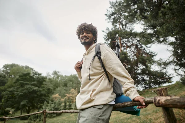 Low angle view of cheerful indian tourist with backpack looking at camera near wooden fence — Stock Photo