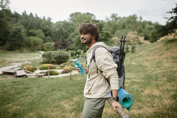 Sorridente jovem turista indiano com mochila segurando garrafa de esportes perto de cerca ao ar livre — Fotografia de Stock