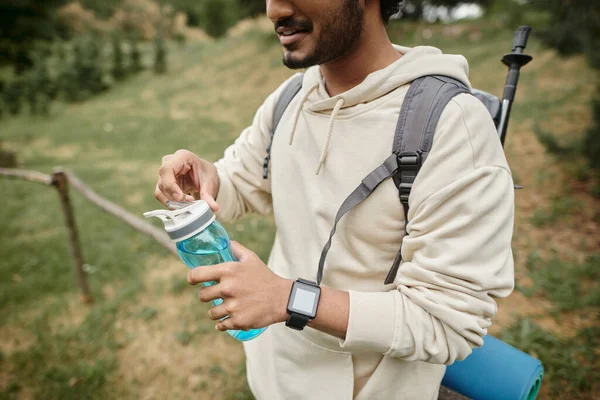 Vista recortada de turista indio con mochila sosteniendo botella deportiva al aire libre - foto de stock