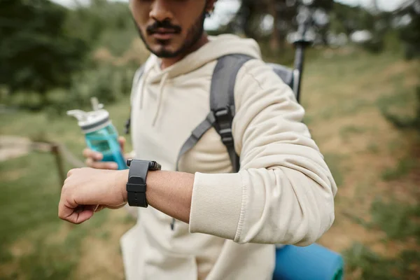 Vista recortada de turista indio con mochila sosteniendo botella deportiva y usando smartwatch al aire libre - foto de stock