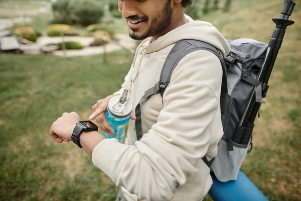 Vista recortada del alegre excursionista indio usando smartwatch y sosteniendo la botella deportiva al aire libre - foto de stock