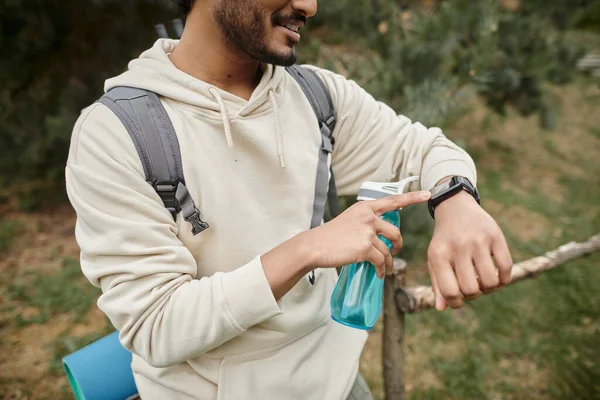 Vista cortada do viajante índio sorridente com mochila e garrafa de esportes usando smartwatch ao ar livre — Fotografia de Stock
