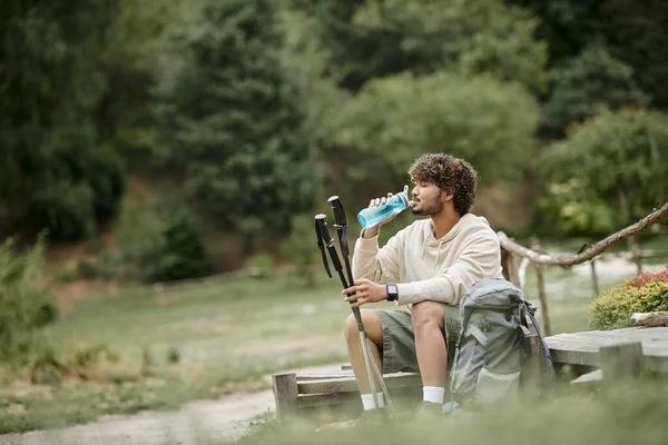 Side view of indian tourist drinking water and holding trekking poles near backpack in forest — Stock Photo