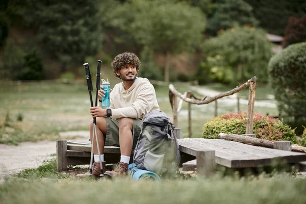 Alegre senderista indio sosteniendo bastones de trekking y botella deportiva cerca de la mochila en el bosque - foto de stock