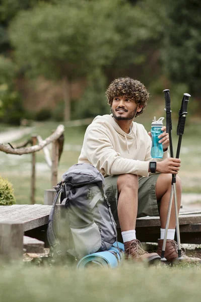 Улыбающийся индийский путешественник держит треккинговые палки и спортивную бутылку возле рюкзака в лесу — стоковое фото
