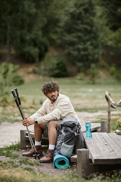 Senderista indio cansado sosteniendo bastones de trekking cerca de la mochila en el bosque borroso - foto de stock