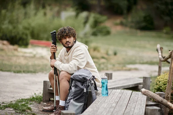 Jeune touriste indien tenant bâtons de trekking près du sac à dos tout en se reposant dans la forêt — Photo de stock