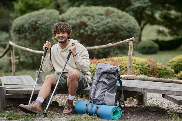 Cheerful indian backpacker holding trekking poles and looking at camera in forest — Stock Photo