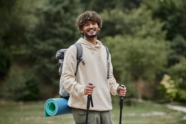 Positive indian backpacker holding trekking poles on path in forest, travel and adventure concept — Stock Photo
