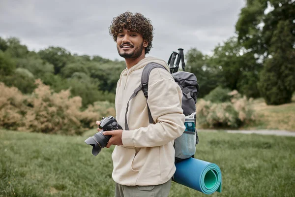 Mochilero indio sonriendo y sosteniendo la cámara digital en el bosque, fotografía y concepto de aventura - foto de stock