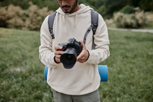 Vue recadrée du photographe indien en sweat à capuche tenant appareil photo professionnel, sac à dos et voyage — Photo de stock
