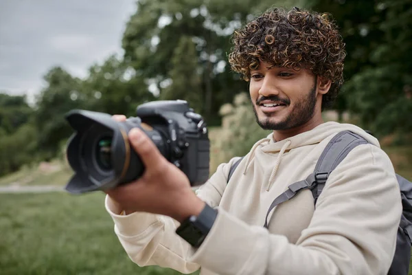 Photographe indien bouclé avec sac à dos prenant des photos sur appareil photo professionnel, aventure et voyage — Photo de stock