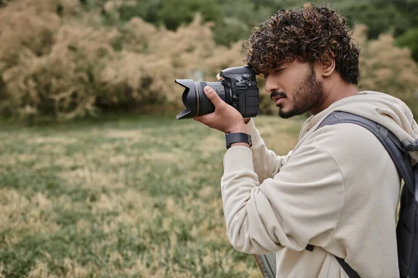 Photographe indien concentré avec sac à dos prenant des photos sur appareil photo professionnel, emplacement naturel — Photo de stock