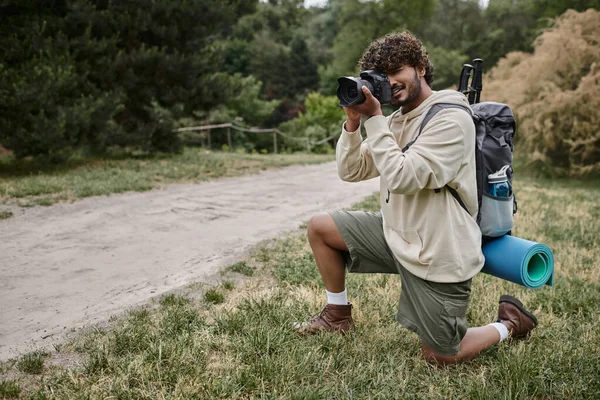 Fotógrafo indiano positivo com mochila tirando foto na câmera profissional, localização natural — Fotografia de Stock