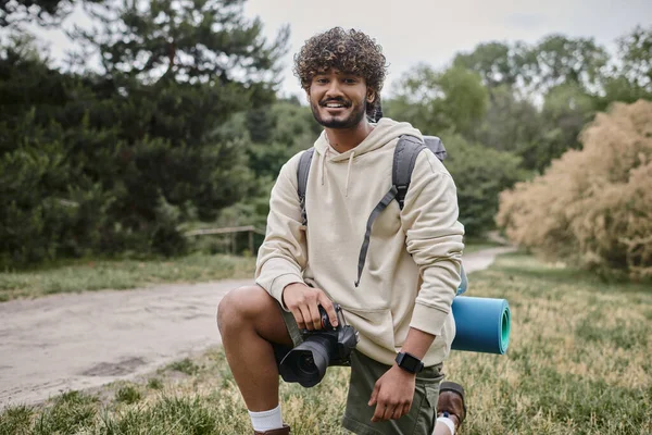Fotógrafo indiano alegre com mochila segurando câmera profissional, localização natural, viajar — Fotografia de Stock