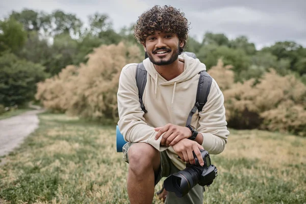 Fotógrafo indio alegre sosteniendo la cámara profesional y mirando la cámara en el bosque durante el viaje - foto de stock