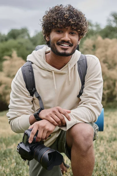 Fotógrafo indio alegre sosteniendo cámara profesional y mirando a la cámara en el bosque, vertical - foto de stock