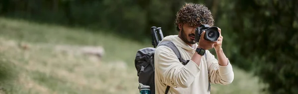Кучерявий індійський турист фотографує на професійній камері, ходить з рюкзаком в лісі, банер — стокове фото