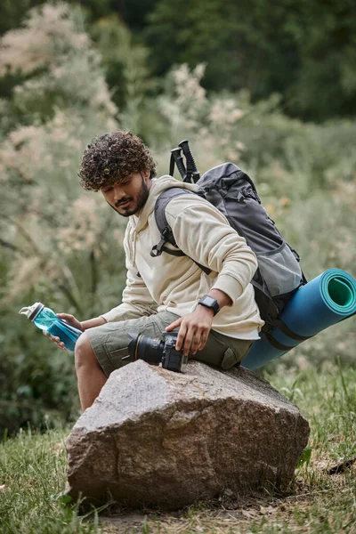Happy indian tourist sitting with backpack on rock and touching camera in forest, adventure — Stock Photo