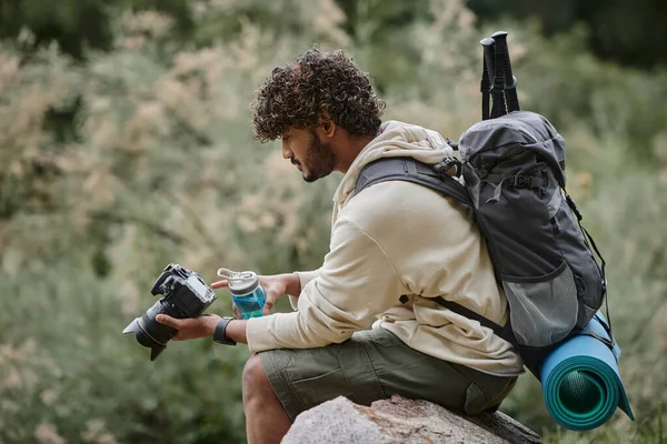 Curly indian tourist holding professional camera and sitting on rock, man with backpack in forest — Stock Photo