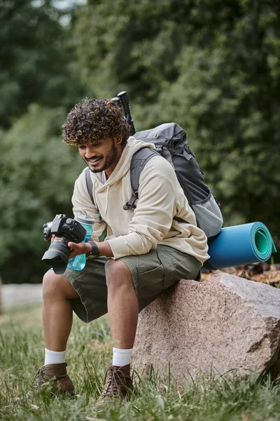 Joyful indian tourist holding professional camera and sitting on rock, man with backpack in forest — Stock Photo