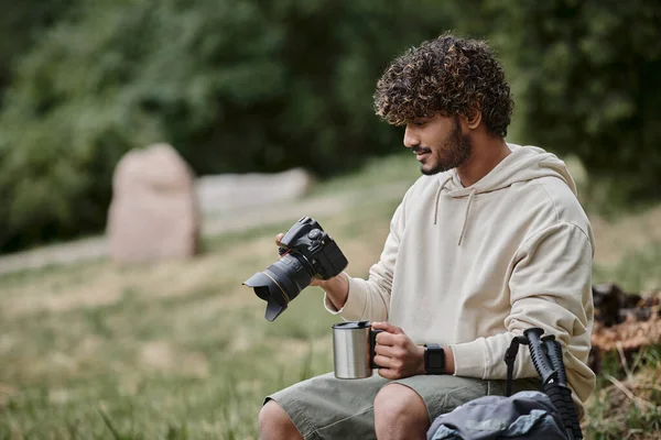 Curly indian tourist holding professional camera and sitting on rock, man with thermos cup in forest — Stock Photo