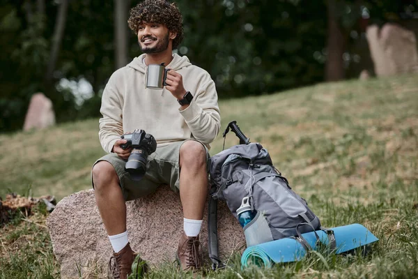 Happy indian tourist holding professional camera and sitting on rock, man with thermos cup in forest — Stock Photo