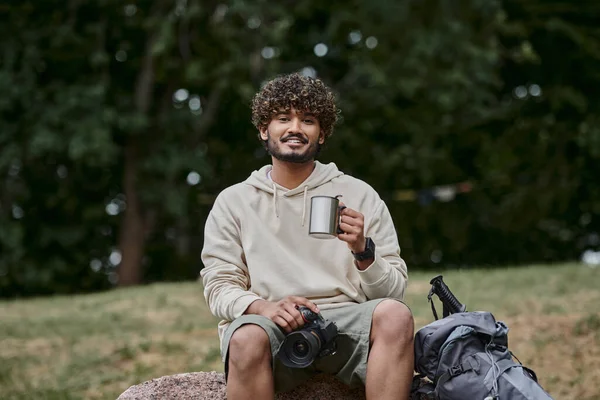 Indian tourist holding professional camera and sitting on rock, happy man with thermos cup in forest — Stock Photo