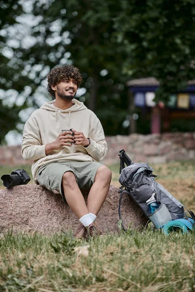 Счастливый индиец держит термос кружку и сидит на скале, кудрявый турист возле туристического снаряжения в лесу — стоковое фото